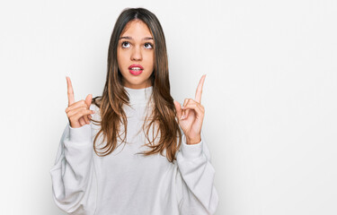 Young brunette woman wearing casual turtleneck sweater amazed and surprised looking up and pointing with fingers and raised arms.