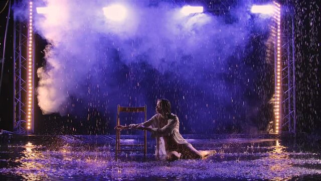 Contemporary modern dance ballerina moves sensually in the rain, sitting on chair. Silhouette of a wet woman in a white shirt move plastically in a dark smoke studio with blue light. Slow motion.