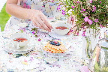 Homemade waffles  with jam and cups with tea on a flowery tablecloth  with woman drinking tea in background outdoors in summertime in the garden