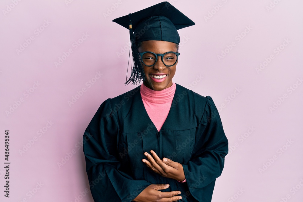 Sticker Young african american girl wearing graduation cap and ceremony robe smiling and laughing hard out loud because funny crazy joke with hands on body.