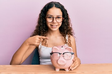 Obraz na płótnie Canvas Teenager hispanic girl holding piggy bank with glasses smiling happy pointing with hand and finger
