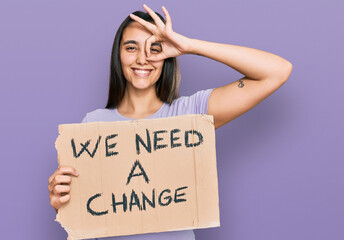 Young hispanic woman holding we need a change banner smiling happy doing ok sign with hand on eye looking through fingers