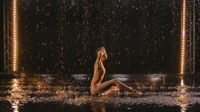 Young woman performs choreography modern ballet with acrobatic elements, bending body on water surface in rain. Silhouette of sexy female in wet swimsuit moves sensually in dark studio. Slow motion.