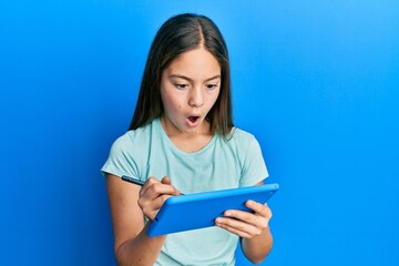 Beautiful brunette little girl using touchpad drawing on screen afraid and shocked with surprise and amazed expression, fear and excited face.