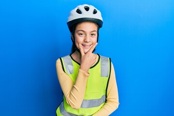 Beautiful brunette little girl wearing bike helmet and reflective vest looking confident at the camera smiling with crossed arms and hand raised on chin. thinking positive.