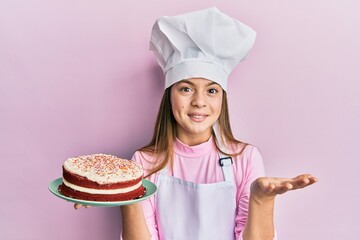 Beautiful brunette little girl wearing baker uniform holding homemade cake celebrating achievement with happy smile and winner expression with raised hand