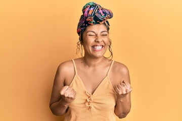 Young hispanic girl wearing hair turban over yellow background excited for success with arms raised and eyes closed celebrating victory smiling. winner concept.