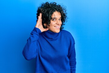 Fototapeta na wymiar Young hispanic woman with curly hair wearing casual winter sweater smiling with hand over ear listening and hearing to rumor or gossip. deafness concept.
