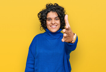 Young hispanic woman with curly hair wearing turtleneck sweater smiling friendly offering handshake as greeting and welcoming. successful business.