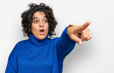 Young hispanic woman with curly hair wearing turtleneck sweater pointing with finger surprised ahead, open mouth amazed expression, something on the front