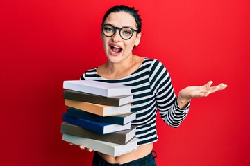 Young caucasian woman wearing glasses and holding books celebrating achievement with happy smile and winner expression with raised hand