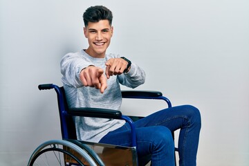 Young hispanic man sitting on wheelchair pointing to you and the camera with fingers, smiling positive and cheerful