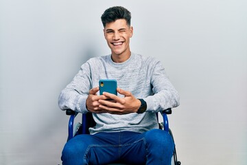 Young hispanic man sitting on wheelchair using smartphone winking looking at the camera with sexy...