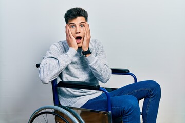 Obraz na płótnie Canvas Young hispanic man sitting on wheelchair afraid and shocked, surprise and amazed expression with hands on face