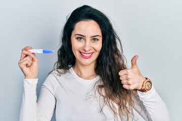 Young hispanic woman holding thermometer smiling happy and positive, thumb up doing excellent and approval sign