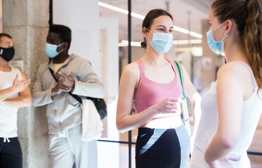 Group of young sporty people in face masks talking at hall at yoga studio, waiting for pilates class - Powered by Adobe