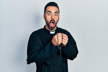 Handsome hispanic priest man with beard praying holding catholic rosary afraid and shocked with...