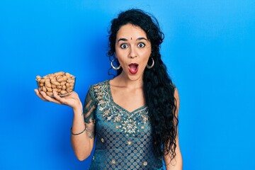 Young woman wearing bindi and traditional kurta holding peanuts scared and amazed with open mouth...