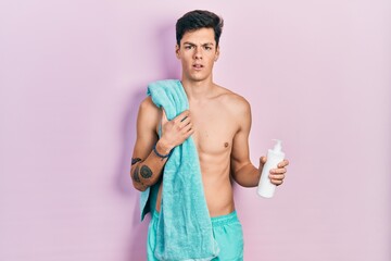 Young hispanic man wearing swimsuit and holding sunscreen lotion in shock face, looking skeptical...