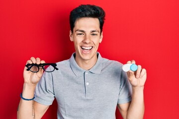 Young hispanic man holding glasses and contact lenses smiling and laughing hard out loud because funny crazy joke.