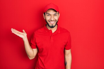 Naklejka premium Hispanic man with beard wearing delivery uniform and cap smiling cheerful presenting and pointing with palm of hand looking at the camera.