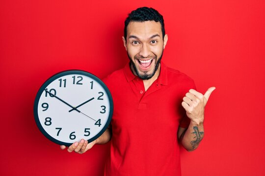 Hispanic man with beard holding big clock pointing thumb up to the side smiling happy with open mouth