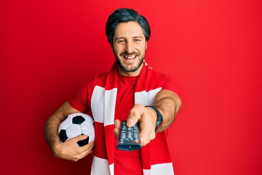 Young Hispanic Man Football Hooligan Holding Ball Using Tv Control Smiling And Laughing Hard Out Loud Because Funny Crazy Joke.