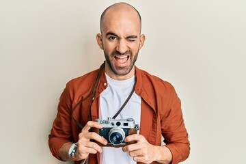 Young hispanic man holding vintage camera winking looking at the camera with sexy expression, cheerful and happy face.