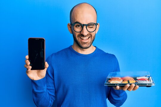 Young hispanic man holding take away food showing smartphone screen winking looking at the camera with sexy expression, cheerful and happy face.