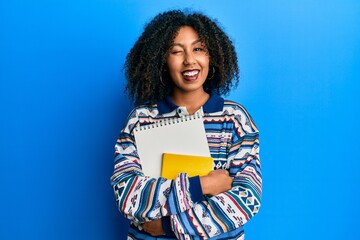 Beautiful african american woman with afro hair holding books winking looking at the camera with...