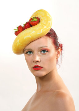 A beautiful young woman poses for the camera. 
She has a yellow python snake on her head.
The python has strawberries wrapped around
its body.