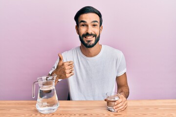 Young hispanic man drinking glass of water sitting on the table smiling happy and positive, thumb up doing excellent and approval sign