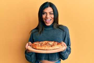 Young latin transsexual transgender woman holding italian pizza winking looking at the camera with...