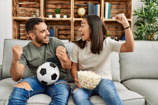 Young Latin Couple Watching Soccer Match Eating Porpcorn At Home.