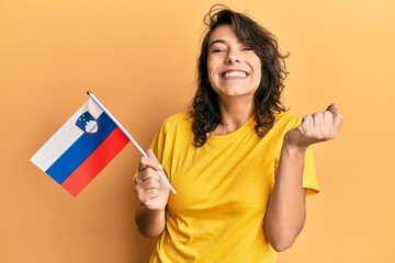 Young hispanic woman holding slovenia flag screaming proud, celebrating victory and success very...