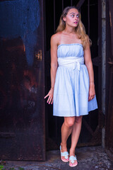 Naklejka premium A pretty blonde woman, dressing in a white-blue dress, white waistband, and sandals, is entering an abandoned house, scared, mysteriously looking around..