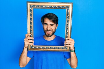Young hispanic man holding empty frame winking looking at the camera with sexy expression, cheerful...