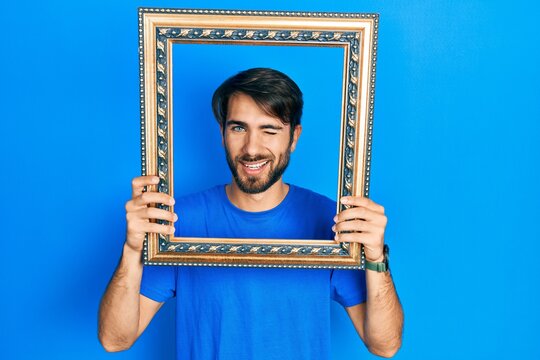 Young hispanic man holding empty frame winking looking at the camera with sexy expression, cheerful and happy face.