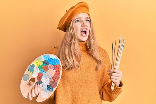 Beautiful young caucasian girl holding paintbrush and painter palette angry and mad screaming frustrated and furious, shouting with anger. rage and aggressive concept.
