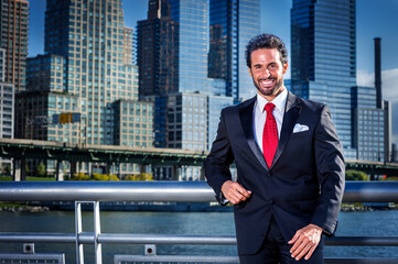 Fototapeta premium Dressing in a black suit and red tie, a handsome, sexy, middle-aged businessman with a beard is standing in the front of a business district, smilingly looking at you..