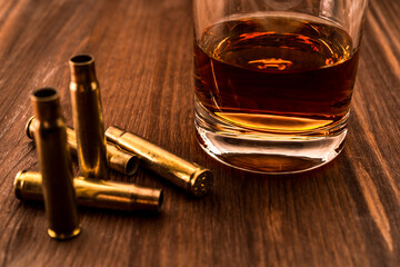 Empty shells from the rifle with glass of whiskey on a wooden table. Focus on the glass of whiskey