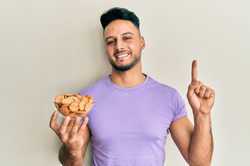 Young arab man holding bowl of salty crackers biscuits smiling with an idea or question pointing finger with happy face, number one