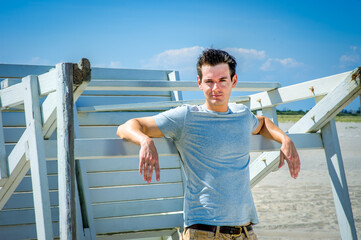 Man waiting for you. Wearing a gray t-shirt and arms resting on a wooden stick, a handsome young man is standing by a wooden structure on the beach, narrowing eyes, charmingly looking at you..