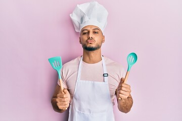 Young arab man wearing professional cook apron and hat holding spoon looking at the camera blowing...
