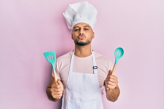 Young arab man wearing professional cook apron and hat holding spoon looking at the camera blowing a kiss being lovely and sexy. love expression.