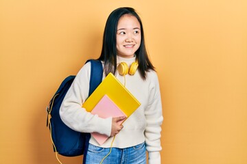 Young chinese girl holding student backpack and books smiling looking to the side and staring away thinking.