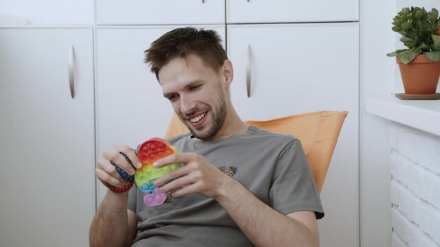 Caucasian man sitting on the balcony, smiling and relieves stress with anti stress toys. In the hands of rainbow pop it and squish ball with slime.