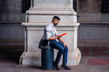 Wearing a gray shirt, blue jeans, black cloth shoes, carrying a shoulder leather bag, a young black man is sitting on a metal pillar on an old street in New York City, relaxing, reading a red book.. - Powered by Adobe