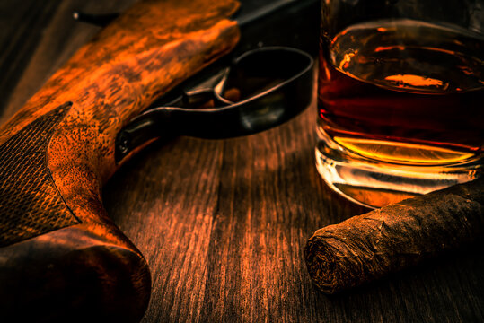 Hunting rifle and glass of whiskey with cuban cigar on the wooden table. Focus on the cuban cigar