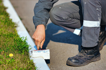A worker in overalls paints a border with white paint on a summer day. Urban services, landscaping. Hand holds a paint brush. Close-up